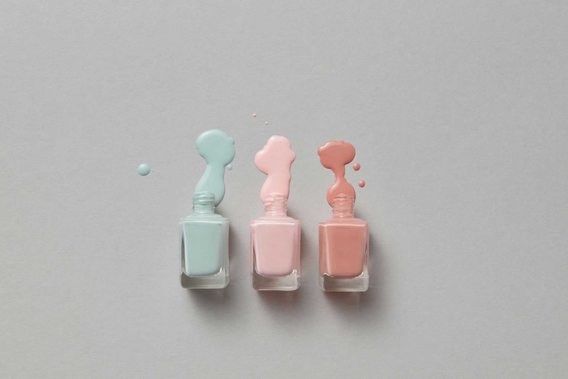 Trois flacons de vernis Ă ongles aux teintes pastel avec des coulures de vernis sur un fond gris neutre.